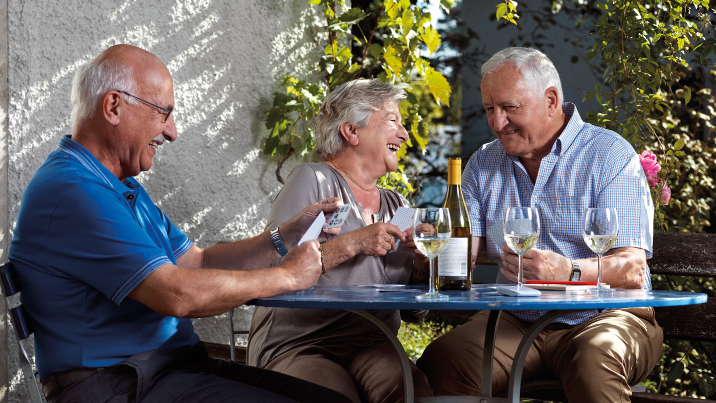 Drei ältere Menschen sitzen am Tisch bei einem Glas Wein und lachen